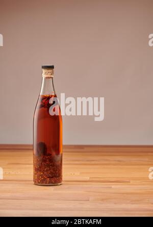 Transparente Flasche hausgemachtes Chilliöl mit Stücken von trockenem Chili, auf einem Holztisch Stockfoto