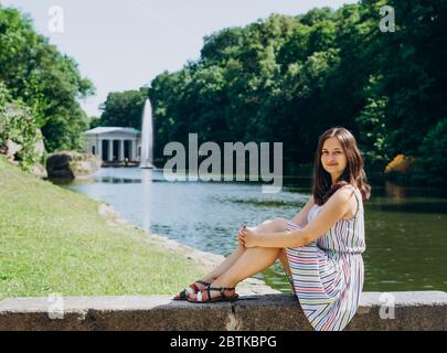 Sofia Park, Uman. Junge Frau auf einer Bank auf dem Hintergrund des Sees mit einem Brunnen. Das Mädchen in einem Kleid sitzt auf einer Steinbank. Stockfoto