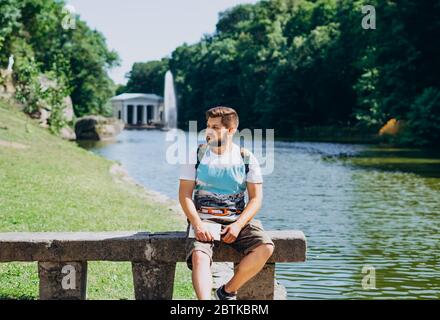 Sofia Park, Uman. Schöner junger Mann sitzt auf einer Bank auf dem Hintergrund des Sees. Männlicher Tourist mit Rucksack auf einer Bank auf der Rückseite sitzend Stockfoto