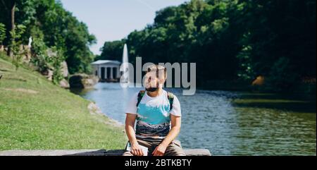 Sofia Park, Uman. Ein schöner Mann mit Rucksack, der auf einer Steinbank im Park sitzt. Junger Mann auf dem Hintergrund des Sees mit einem hohen Brunnen. A m Stockfoto