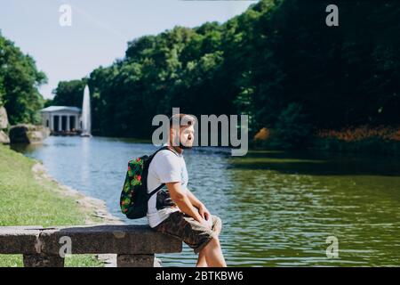 Sofia Park, Uman. Ein schöner Mann mit Rucksack, der auf einer Steinbank im Park sitzt. Junger Mann auf dem Hintergrund des Sees mit einem hohen Brunnen. A m Stockfoto