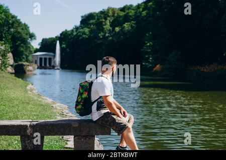 Sofia Park, Uman. Ein schöner Mann mit Rucksack, der auf einer Steinbank im Park sitzt. Junger Mann auf dem Hintergrund des Sees mit einem hohen Brunnen. A m Stockfoto