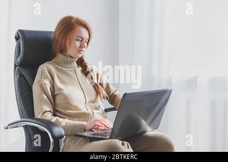 Junge kaukasische Frau sitzt in Ledersessel mit Laptop arbeiten von zu Hause, freiberufliche, soziale Distanzierung und Remote-Arbeit-Konzept. Stockfoto