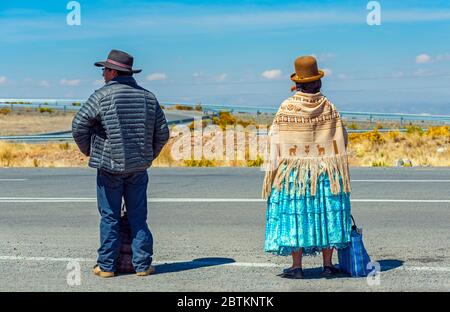 Ein paar Bolivianer Aymara Indigene, mit der Frau in traditioneller Kleidung, warten auf den Transport entlang einer Autobahn, La Paz, Bolivien. Stockfoto