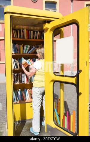 21. Mai 2020, Sachsen-Anhalt, Wörlitz: In einer alten Telefonzelle steht eine Frau an einem Regal mit Büchern, die sie kostenlos mitnehmen können. Mit der Aufschrift 'Befreite Bücher' gehört die stillgesetzte Telefonzelle zur Antiquariatsbuchhandlung 'Schiefes Haus' in Wörlitz. Auf einem Schild an der Tür werden die Interessenten begrüßt und aufgefordert, die Bücher gerade zu stellen. Bücher, die mitgebracht werden, können sortiert oder in der Antiquariatsbuchhandlung abgegeben werden. Foto: Waltraud Grubitzsch/dpa-Zentralbild/ZB Stockfoto