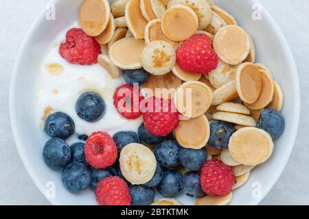 Schüssel mit hausgemachten Mini-Müsli Pfannkuchen mit Heidelbeeren Himbeeren Joghurt und Ahornsirup Stockfoto