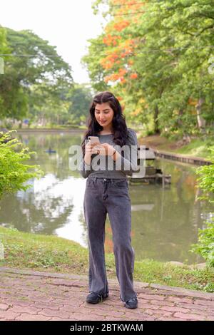 Ganzkörper-Aufnahme von jungen schönen indischen Frau mit Telefon im Park im Freien Stockfoto