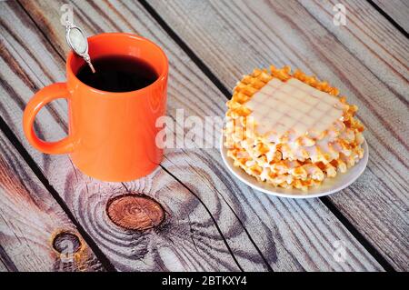 Eine orangefarbene Tasse schwarzen Kaffee mit einem Löffel und einem Teller mit einem Stapel Pariser Waffeln mit Sirup. Nahaufnahme. Stockfoto