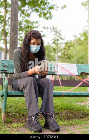 Junge indische Frau mit Maske mit Telefon während des Sitzens mit Abstand auf Parkbank Stockfoto