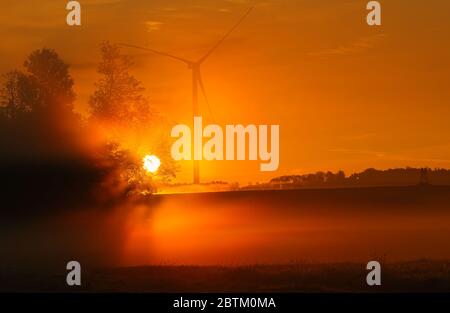 Dummerstorf, Deutschland. Mai 2020. Nebel liegt über einer Wiese, während die Sonne hinter einer Windturbine aufgeht. Quelle: Jens Büttner/dpa-Zentralbild/dpa/Alamy Live News Stockfoto