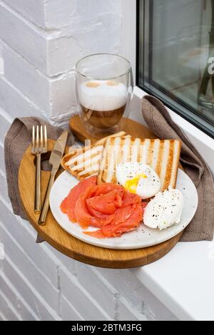 Geröstetes Brot, gesalzener Lachs und pochiertes Ei mit Frischkäse und einer Tasse Latte Macciato auf weißem Fensterbrett. Stockfoto