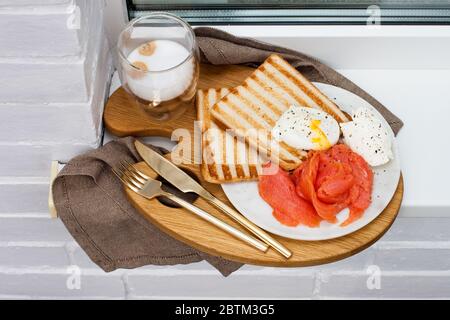 Geröstetes Brot, gesalzener Lachs und pochiertes Ei mit Frischkäse und einer Tasse Latte Macciato auf weißem Fensterbrett. Stockfoto