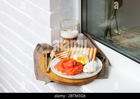 Geröstetes Brot, gesalzener Lachs und pochiertes Ei mit Frischkäse und einer Tasse Latte Macciato auf weißem Fensterbrett. Stockfoto