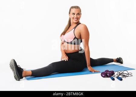 Junge fröhliche Frau mit Übergewicht in sportlichem Oberteil und Leggings Stretching auf Yoga-Matte, während glücklich beiseite über weißen Hintergrund Stockfoto