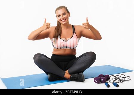 Junge lächelnde Frau mit Übergewicht in sportlichem Oberteil und Leggings sitzt auf Yoga-Matte und zeigt glücklich große Daumen nach oben in der Kamera über weiß Stockfoto