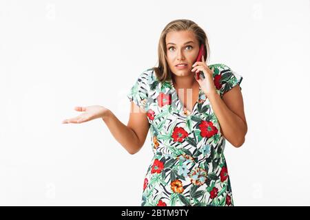 Junge Frau in bunten Kleid erstaunlich in der Kamera zu suchen, während auf Handy über weißem Hintergrund sprechen Stockfoto