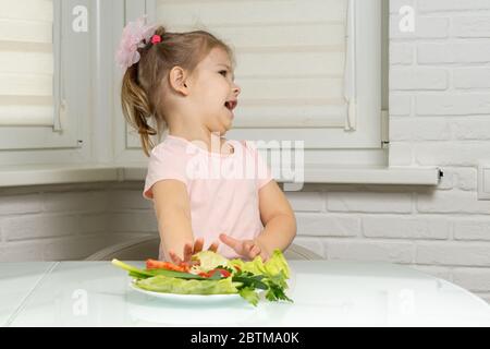 Ein kleines Mädchen sitzt an einem Tisch in der Küche und drückt weg einen Teller Gemüse. Kinder wollen nicht Gemüse essen Stockfoto