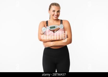 Junge lächelnde Frau mit Übergewicht in sportlichem Oberteil hält Waage in den Händen, während glücklich in der Kamera über weißem Hintergrund schauen Stockfoto
