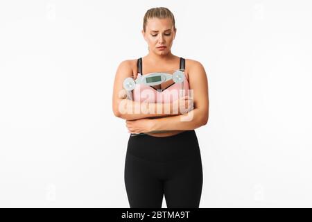 Junge verärgert Frau mit Übergewicht in sportlichen Top-Holding-Waage in den Händen über weißem Hintergrund isoliert Stockfoto