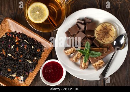 Duftender Blumentee mit Zitrone und Zimt, ein Teller mit Süßigkeiten: Schokolade, Honig und Minze, und trockene Teeblätter in einem Teller auf natürlichem Hintergrund. Stockfoto