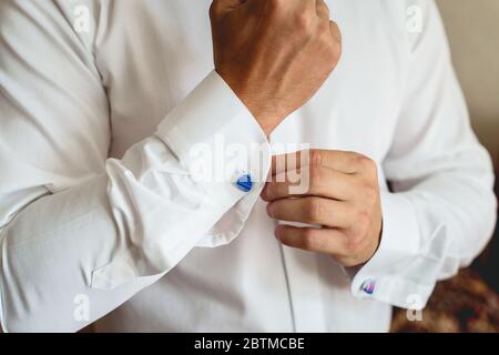 Ein Mann befestigt Manschettenknöpfe auf dem weißen Hemd. Nahaufnahme einer Männerhand mit einem weißen Hemd und Manschettenknöpfen. Stockfoto