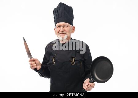 Alter fröhlicher Mann mit grauem Bart in schwarzer Schürze und Kappe hält Pfanne und Messer in den Händen, während glücklich in der Kamera über weißem Hintergrund zu sehen Stockfoto