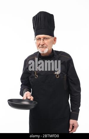 Alter schöner Mann mit grauem Bart in schwarzer Schürze und Kappe hält Pfanne in der Hand, während nachdenklich in der Kamera über weißem Hintergrund Stockfoto