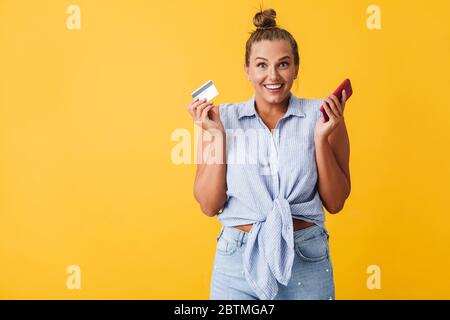 Fröhliche Frau im Hemd freudig in der Kamera suchen, während Kreditkarten und Handy in den Händen über gelben Hintergrund halten Stockfoto