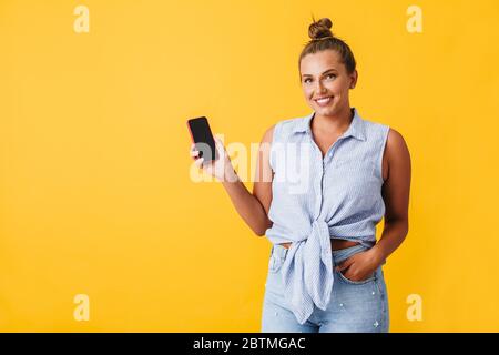 Schöne lächelnde Frau im Hemd glücklich in der Kamera suchen, während neue Handy über gelben Hintergrund zeigen Stockfoto