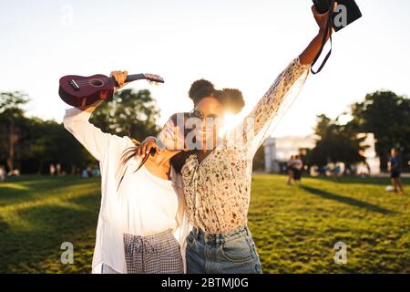 Ziemlich lächelnde Mädchen mit wenig Gitarre und polaroid Kamera glücklich ihre Hände hoch, während sie Zeit zusammen im Park verbringen Stockfoto