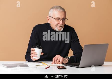 Alter schöner Mann mit grauen Haaren und Bart in Brillen und Pullover sitzt am Tisch nachdenklich arbeiten auf Laptop mit Kaffee in der Hand zu gehen über Stockfoto