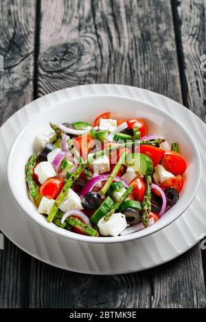 Feta-Salat mit grünem gegrilltem Spargel, Kirschtomaten, frischen Gurken, schwarzen Oliven, roten Zwiebelringen mit Olivenöl auf einer weißen Schüssel auf einem ru serviert Stockfoto