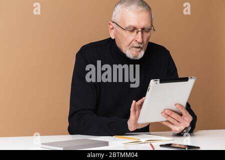 Alter schöner Mann mit grauen Haaren und Bart in Brillen und Pullover sitzt am Tisch nachdenklich mit der Tablette über beigefarbenem Hintergrund isoliert Stockfoto