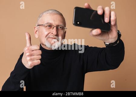 Alter schöner Mann mit grauen Haaren und Bart in Brillen und Pullover glücklich zeigen Daumen bis Geste während der Aufnahme auf Handy über beige Stockfoto