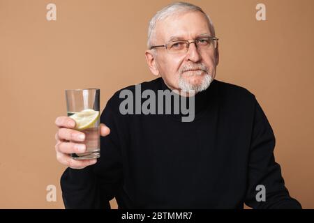 Alter schöner Mann mit grauen Haaren und Bart in Brillen und Pullover nachdenklich in der Kamera halten Glas von reinem Wasser mit einer Scheibe Zitrone Stockfoto