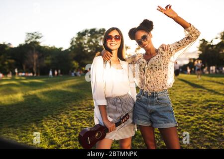 Schöne lächelnde Mädchen in Sonnenbrille mit wenig Gitarre glücklich in der Kamera suchen, während die Zeit zusammen im Park Stockfoto