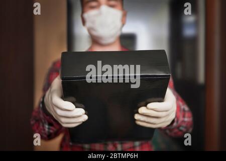Nach Hause liefern, Online-Bestellung. Mann in einer medizinischen Maske und Gummihandschuhe mit schwarzer Box, ein Paket in den Händen. Lieferung während der Quarantäne der Stockfoto
