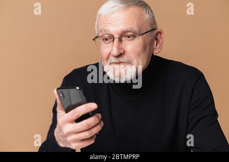 Alt attraktiven Mann mit grauen Haaren und Bart in Brillen und Pullover erstaunlich auf Handy über beigefarbenen Hintergrund isoliert suchen Stockfoto