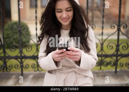 Lächelndes schwarzhaariges Mädchen, das auf der Straße ist und ein Mobiltelefon benutzt Stockfoto