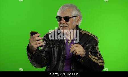 Ältere stilvolle Großvater kaukasischen reifen Rocker und Biker Mann mit App auf dem Smartphone für Videoanruf. Hintergrund der Chroma-Taste. Alte ältere Großeltern in Lederjacke Surfen auf dem Handy Stockfoto