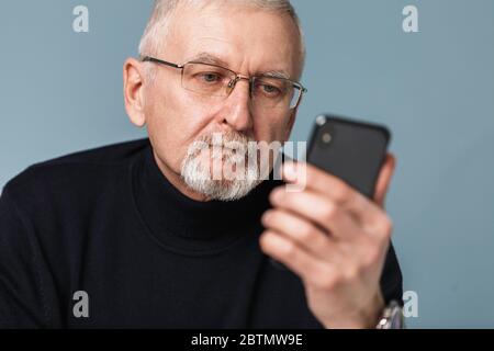 Alte nachdenkliche Mann mit grauen Haaren und Bart in Brillen und Pullover nachdenklich mit Handy über blauem Hintergrund isoliert Stockfoto