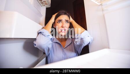 Verzweifelte Frau, die zu Hause auf den leeren Kühlschrank schaut Stockfoto