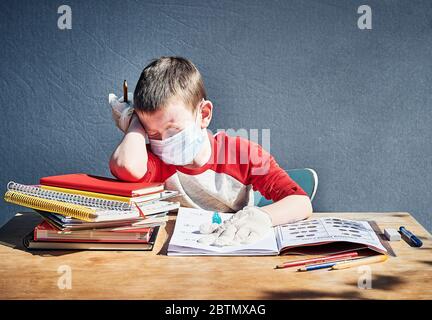 Der fünfjährige Junge sitzt mit Büchern an einem Schulschreibtisch und trägt eine Gesichtsmaske und Handschuhe (PSA). Stockfoto