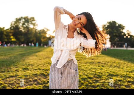 Hübsches lächelndes Mädchen in weißem Hemd, das beim Tanzen im Stadtpark freudig zur Seite schaut Stockfoto