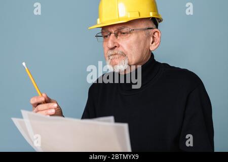 Alte schöne ernst Bussinesman mit grauen Bart in Brillen und gelben Schutzhelm nachdenklich Blick auf Skizze mit Bleistift in der Hand über blau Stockfoto