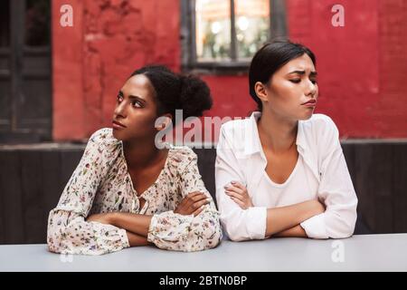 Zwei verärgerte Mädchen beleidigten sich über die anderen, die traurig beiseite schauten, während sie Zeit zusammen im Innenhof des Cafés verbrachten Stockfoto