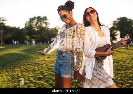 Fröhliche Mädchen in Sonnenbrille glücklich spielen auf wenig Gitarre und tanzen, während Sie Zeit zusammen im Stadtpark verbringen Stockfoto