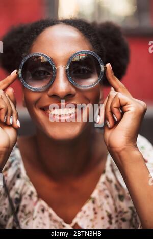 Porträt von fröhlichen afrikanischen Mädchen mit dunklen lockigen Haaren in Sonnenbrille freudig in der Kamera suchen Stockfoto