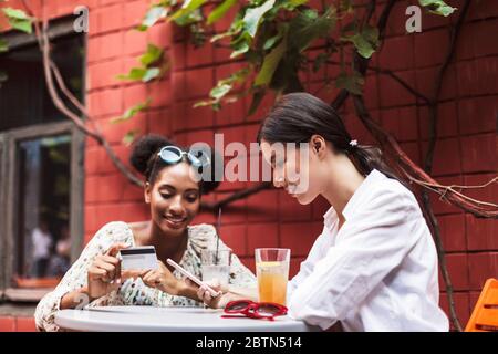 Zwei recht lächelnde Mädchen, die Handy und Kreditkarte benutzen, während sie gerne Zeit zusammen im gemütlichen Innenhof des Cafés verbringen
