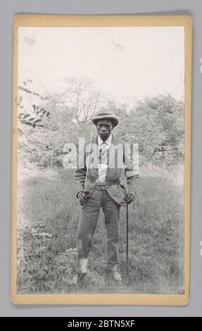 Foto eines unbekannten Mannes, der einen Säbel hält. Dieser fotografische Druck zeigt einen nicht identifizierten Mann, der in einem bewaldeten Feld steht und einen karierten dreiteiligen Anzug, ein gestreiftes Hemd, einen Krawatte mit Polka-Punkten, einen Glockenhut und eine ovale US-Gürtelplatte auf einem Ledergürtel mit Scheide trägt. Stockfoto